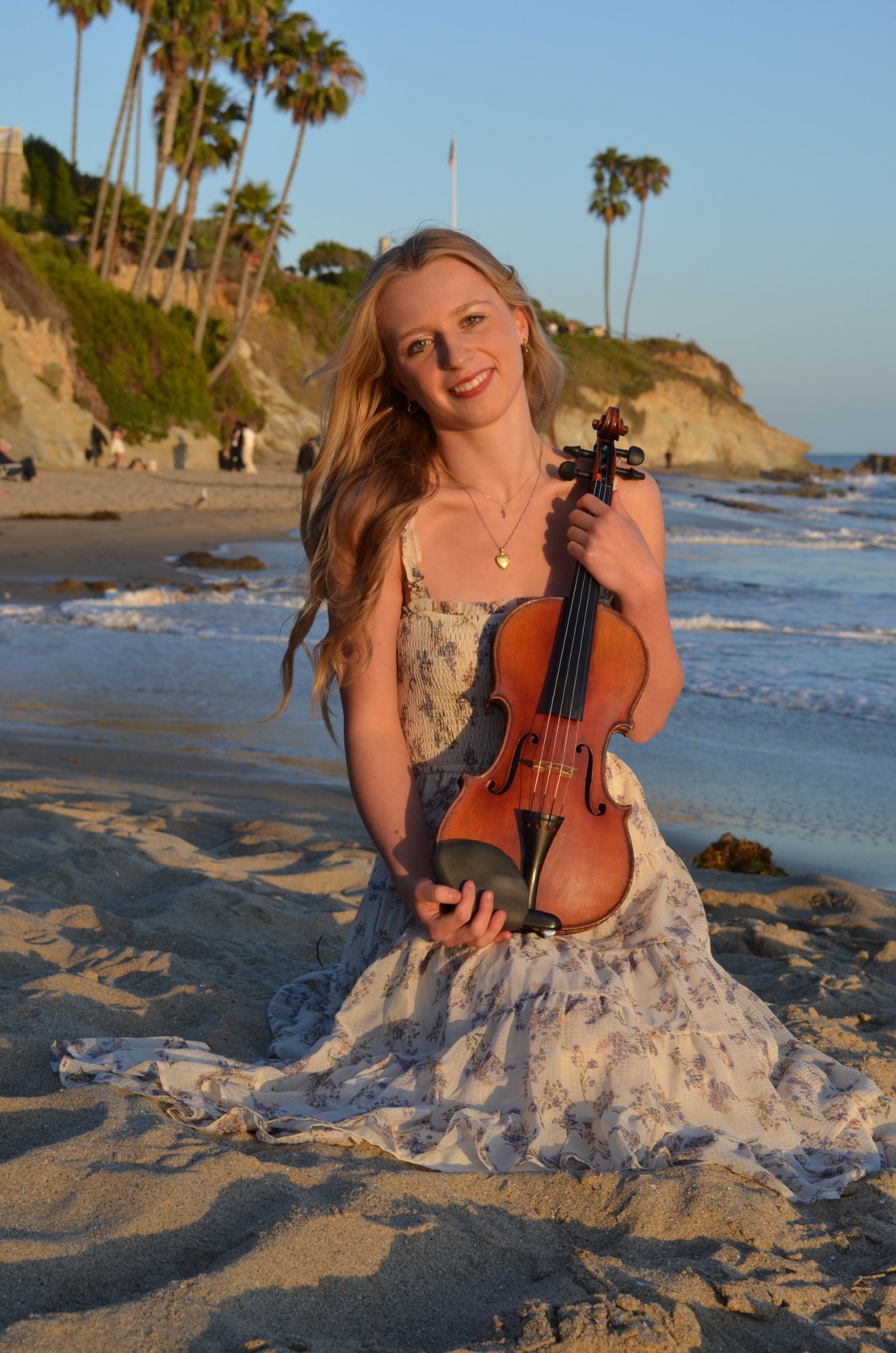 Hannah smiling on the beach at sunset in a floral dress, holding her violin